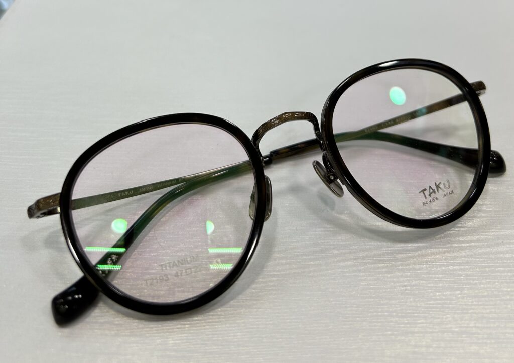 Round, black-framed eyeglasses with tinted lenses lie on a white surface. The left lens has "TITANIUM" and "24-0189 47[]21" etched on it, while the right bears the brand name "TAKU." These exquisite 眼鏡 are the perfect choice for those seeking both style and durability.